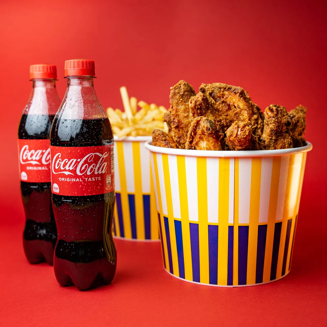 Kanta pohanih pilećih krilaca i pomfrita uz dvije boce Coca-Cole na crvenoj pozadini